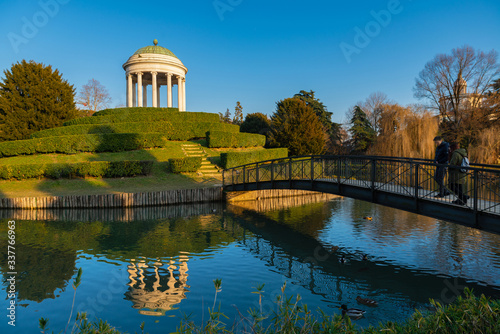 A little monopteros ionic style temple is reflected on the lake of Parco Querini, the main public park of Vicenza city. A sunny and cloudless winter day. Couples of mallards swim quiet on water.
