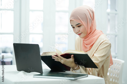 Asian muslim high school student girl reading the book while doing a educational project in front of laptop screen. learning, studying, technology and education concept. 