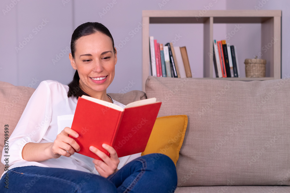 Pretty young brunette woman smiling reading a book sitting on the sofa at home. She is wearing a white shirt and a ponytail. Pretty young brunette woman smiling reading a book sitting on the sofa at home. She is wearing a white shirt and a ponytail.