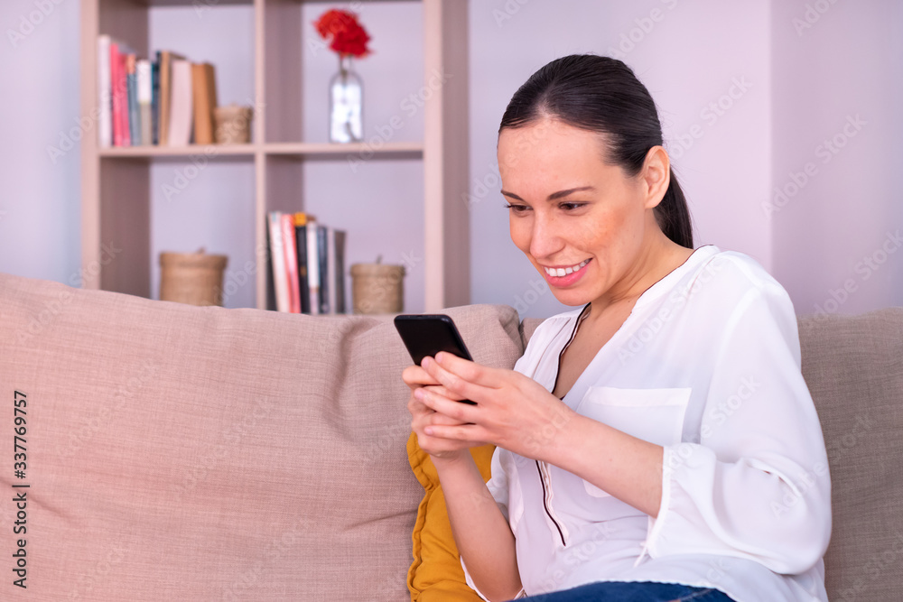 Pretty young brunette woman smiling writes a message on the smartphone sitting on the sofa at home. She is wearing a white shirt and a ponytail. Pretty young brunette woman smiling writes a message on the smartphone sitting on the sofa at home. She is wearing a white shirt and a ponytail.