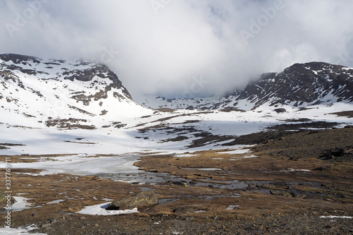The mountain valley with the small river, dark rocky slopes covered by white snow, the blue sky with white clouds.