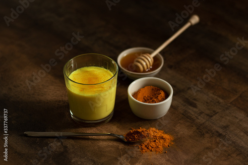 Turmeric Milk is poured into a glass. Next to it are the ingredients for making a drink - milk in a bottle on a napkin, spices and honey. Golden drink on dark moody background, selective focus. Top