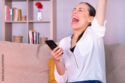 Beautiful young brunette woman euphoric receives good news by the smartphone sitting on the sofa at home. She is wearing a white shirt and a ponytail.