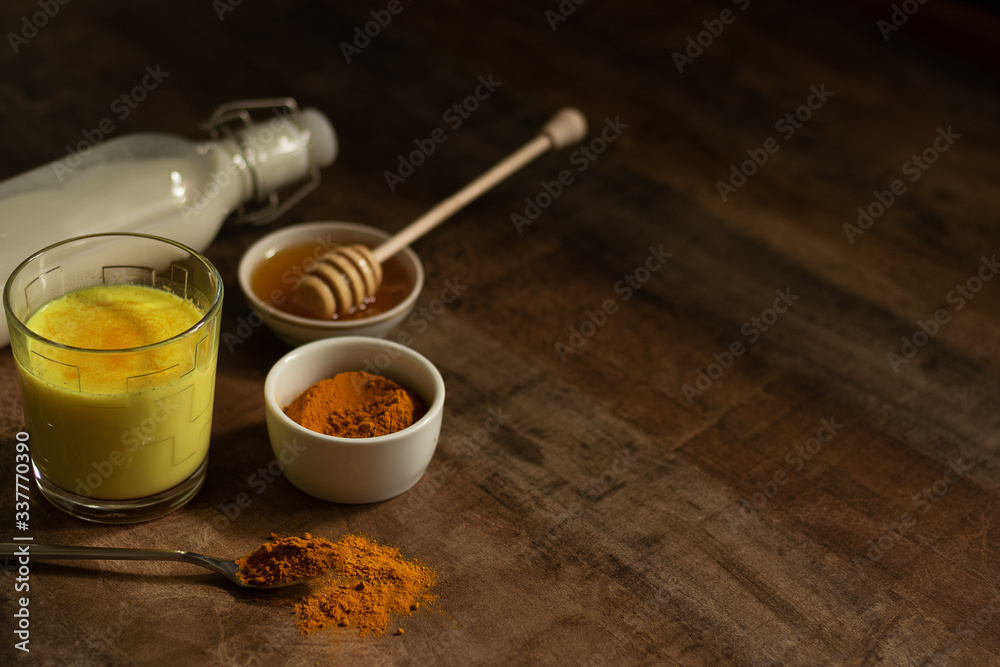 Turmeric Milk is poured into a glass. Next to it are the ingredients for making a drink - milk in a bottle on a napkin, spices and honey. Golden drink on dark moody background, selective focus. Top Turmeric Milk is poured into a glass. Next to it are the ingredients for making a drink - milk in a bottle on a napkin, spices and honey. Golden drink on dark moody background, selective focus. Top