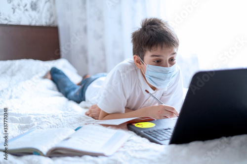 the boy sits at home on the bed in a sterile mask and undergoes online training on a laptop. Distance learning, online lessons. Quarantine and self-isolation during the school year