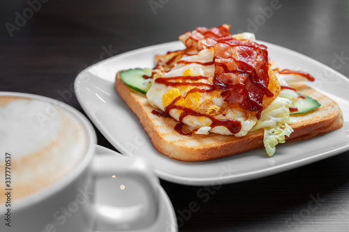 An egg and bacon sandwich on a white plate next to a cappuccino on a dark wooden table.