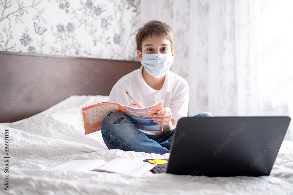 the boy sits at home on the bed in a sterile mask and undergoes online training on a laptop. Distance learning, online lessons. Quarantine and self-isolation during the school year