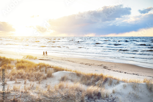 Fototapeta Naklejka Na Ścianę i Meble -  A couple is walking through the Baltic sea shore at sunset. Sand dunes and plants close-up. Colorful evening clouds. Waves and water splashes. Idyllic seascape. Latvia