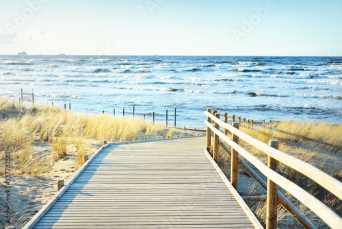 Fototapeta Naklejka Na Ścianę i Meble -  A wooden pathway to the Baltic sea at sunset. Sand dunes and plants in the background. Clear blue evening sky. Idyllic seascape. Latvia