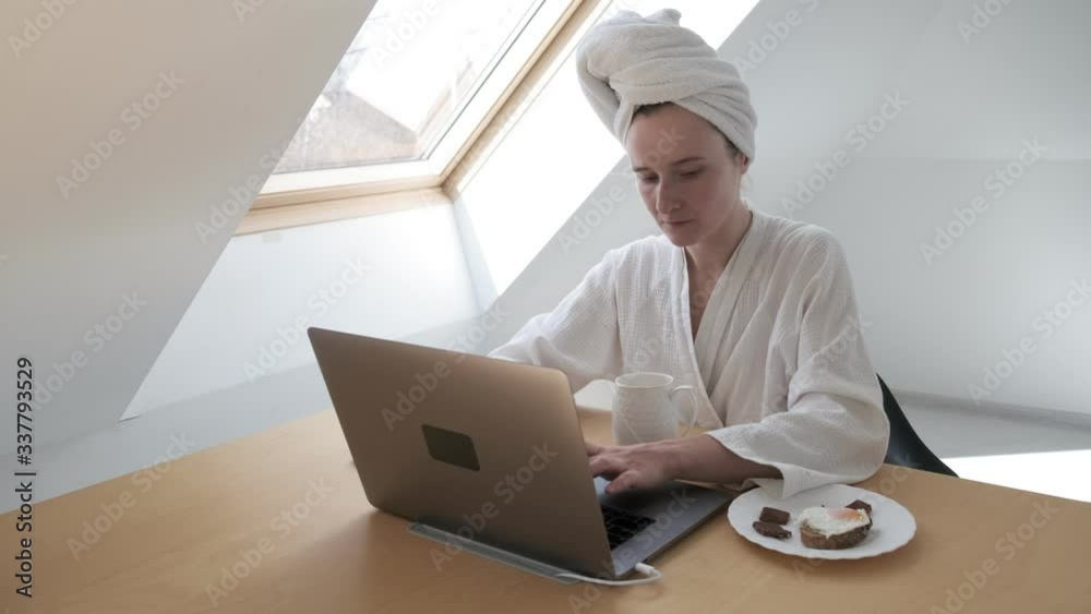 young freelance woman in white bathrobe, towel on head during telework due to quarantine of coronovirus pandemic COVID-2019. Girl eating sandwich, having breakfast while distant working at home laptop young freelance woman in white bathrobe, towel on head during telework due to quarantine of coronovirus pandemic COVID-2019. Girl eating sandwich, having breakfast while distant working at home laptop