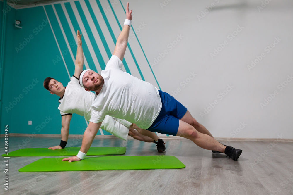 Fat man is working hard in the fitness gym with his personal trainer doing exercises on the mat at the limit of possibilities