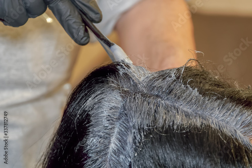 Hair dye color applied on the scalp by a hairdresser wearing black rubber gloves. At-home hair coloring styling with a solid level of black paint mix coverage, using a brush on the hair roots.