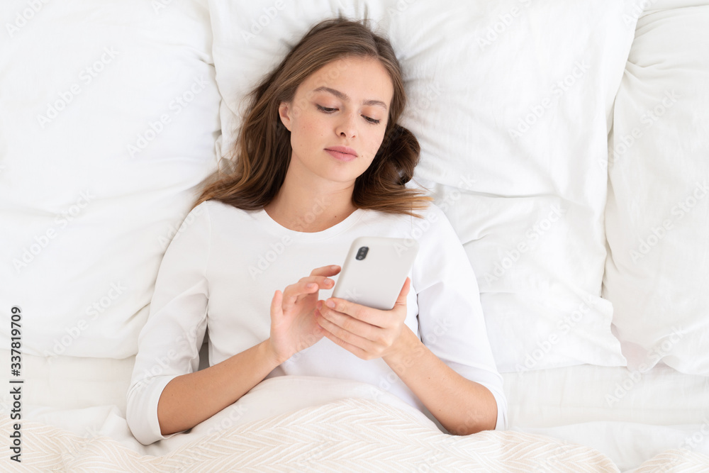 Young woman on white bed in morning holding smartphone and scrolling newsfeed of social network with fingers, reading news, looking serious Young woman on white bed in morning holding smartphone and scrolling newsfeed of social network with fingers, reading news, looking serious