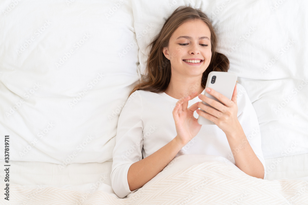 Young girl smiling and laughing at what she sees on her smartphone screen after awakening in morning, surrounded by white pillows Young girl smiling and laughing at what she sees on her smartphone screen after awakening in morning, surrounded by white pillows