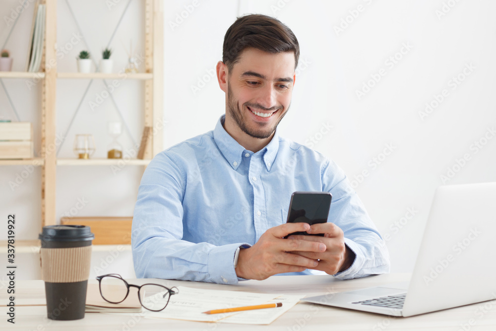 Business guy dressed in blue shirt sitting at desk in his office, holding phone and smiling happily while reading funny content on screen Business guy dressed in blue shirt sitting at desk in his office, holding phone and smiling happily while reading funny content on screen