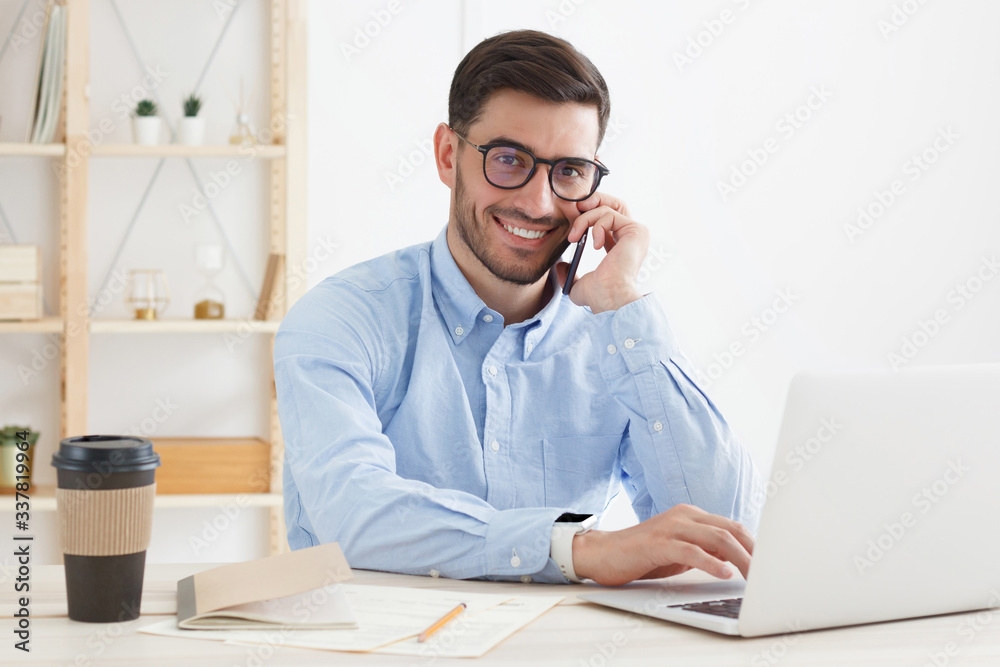 Guy in blue shirt and eyeglasses talking on phone during his working day, solving business tasks, filling forms on laptop, smiling happily at camera