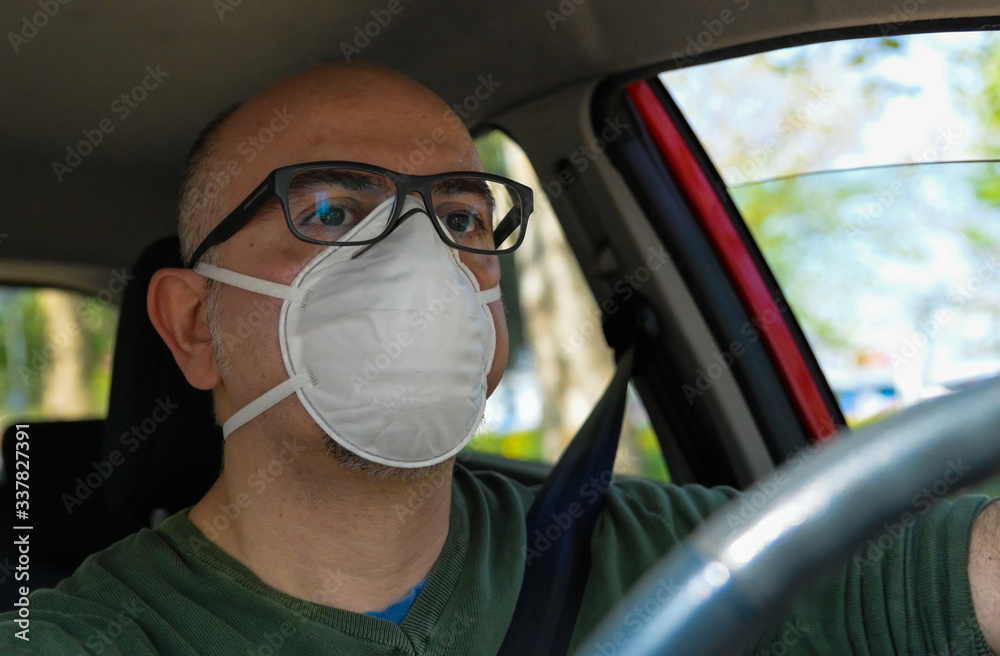 Turin, Piedmont, Italy. April 2020. Coronavirus pandemic: portrait of a Caucasian man driving the car wearing a white mask to avoid contagion. Selective focus on man and blurred background. Turin, Piedmont, Italy. April 2020. Coronavirus pandemic: portrait of a Caucasian man driving the car wearing a white mask to avoid contagion. Selective focus on man and blurred background.