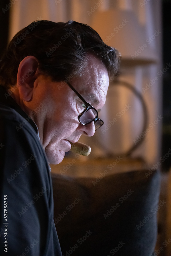 Close-up side profile of a middle aged man looking down through his reading glasses with a cigar clenched in his mouth. The dark room is moodily lit by the light of the television Close-up side profile of a middle aged man looking down through his reading glasses with a cigar clenched in his mouth. The dark room is moodily lit by the light of the television