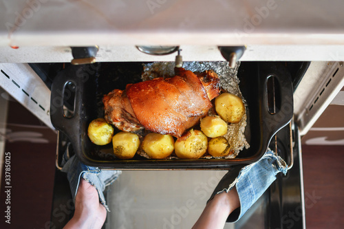 Woman hands taking out roasted pork foreshank with potatoes out of the oven. Top view. Selective focus. photo