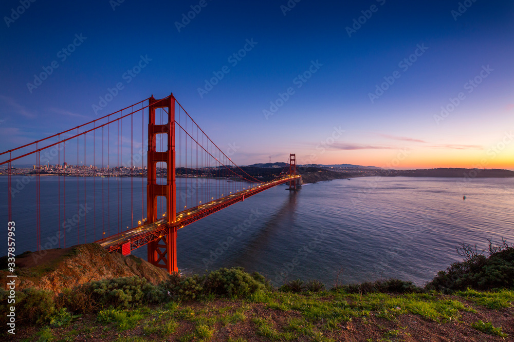 Obraz premium Sunset over the Golden Gate Bridge with the skyline of San Francisco, California in the background.