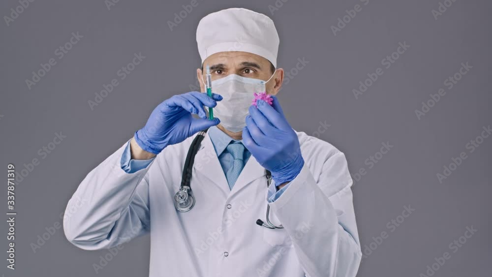 Male middle aged doctor in medical uniform and mask holds syringe, injects test vaccine in covid-19 virus cell, grey background. Coronavirus vaccine research, injection, medicine, vaccination concept Male middle aged doctor in medical uniform and mask holds syringe, injects test vaccine in covid-19 virus cell, grey background. Coronavirus vaccine research, injection, medicine, vaccination concept