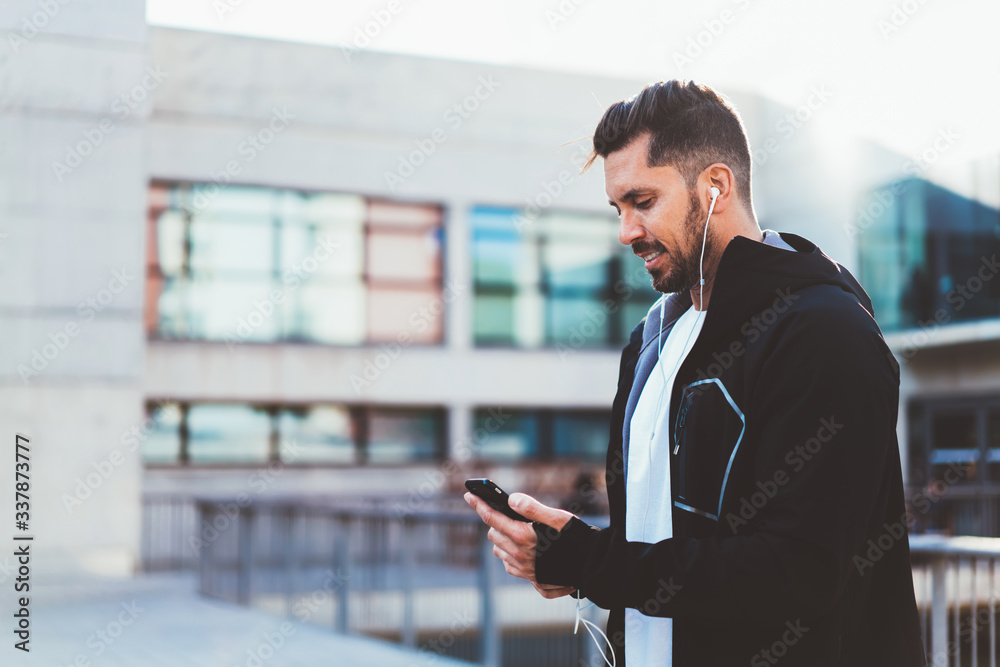Positive caucasian 20s male i sports jacket using app for tracking results for workout on mobile phone, smiling man jogger prepare for cardio training using cellular on street for listening songs Positive caucasian 20s male i sports jacket using app for tracking results for workout on mobile phone, smiling man jogger prepare for cardio training using cellular on street for listening songs