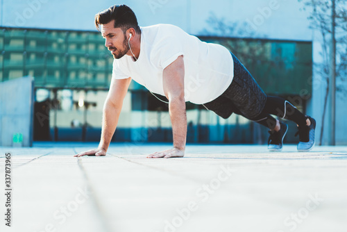Young guy doing pushing from the floor outdoors training flexibility and strength reaching vitality goals, sportsman having morning workout doing exercises in earphones having physical power