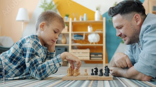 Smart child playing chess with dad at home lying on floor together having fun with intellectual sports game. Happy family and leisure activity concept.