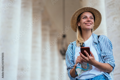 Cheerful Caucasian hipster girl with modern smartphone technology and navigation map in hands smiling during recreation Italian vacations, happy traveller with mobile device enjoying getaway journey © BullRun