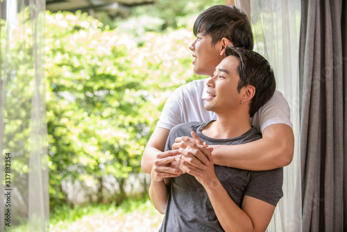 Sweet young Asian homosexual gay man couple hugging and looking to the garden at home. Handsome guy LGBTQ lover enjoy romantic lifestyle together. Same-sex marriage and LGBT relationship concept.