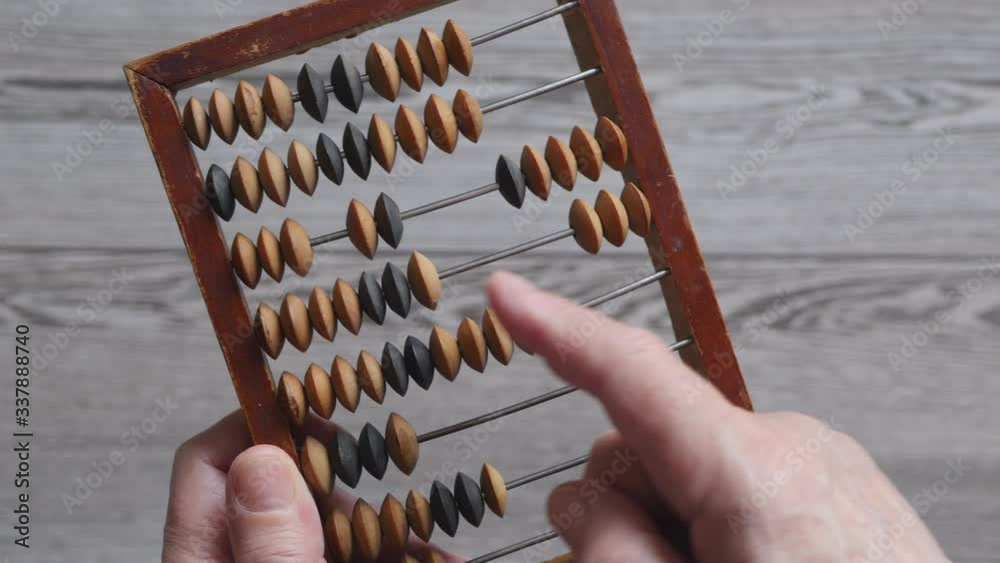 A Elderly Man Is Holding And Moves Wooden Knuckles On A Outdated Vintage Wooden Abacus On A Gray Background, Close-up On The Left Side. A Elderly Man Is Holding And Moves Wooden Knuckles On A Outdated Vintage Wooden Abacus On A Gray Background, Close-up On The Left Side.