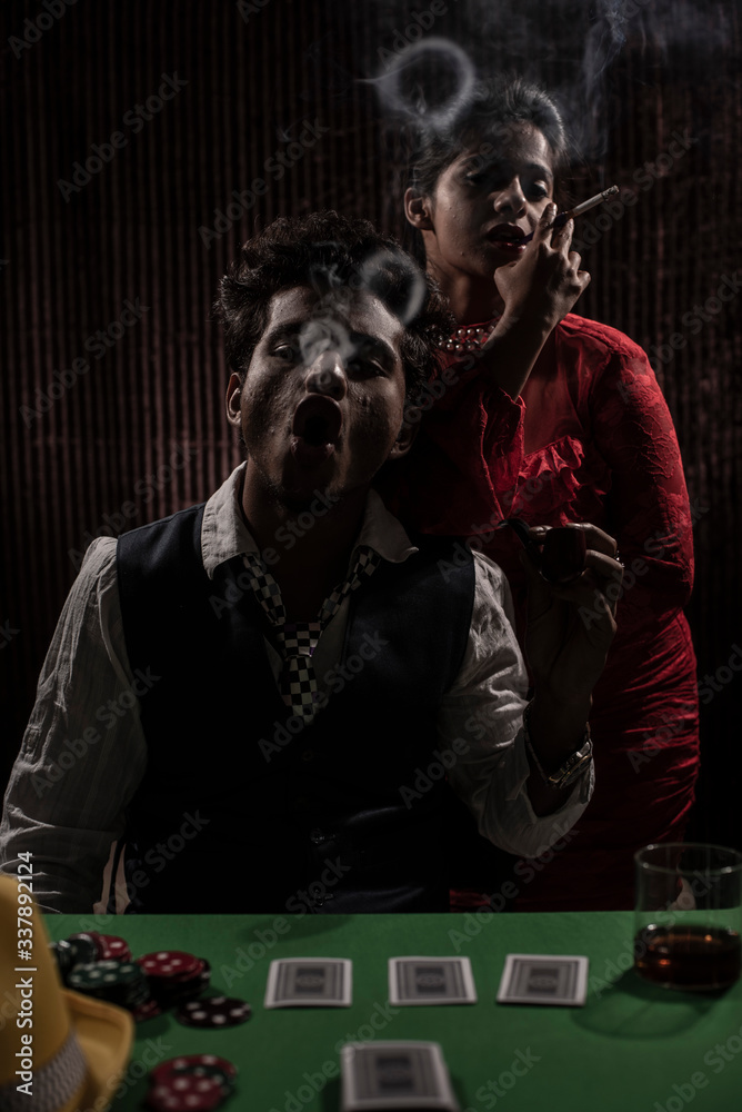An young and attractive Indian brunette couple in vintage suits smoking in a casino poker table in brown textured copy space background. Gambling and addictive lifestyle. An young and attractive Indian brunette couple in vintage suits smoking in a casino poker table in brown textured copy space background. Gambling and addictive lifestyle.