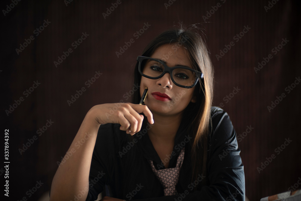 Portrait of an young Indian brunette girl in black corporate suit with a pen in dark copy space studio background. Indian lifestyle and corporate 
