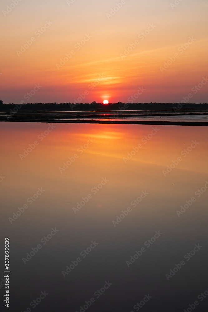 Fire and marmalade colors of sunrise light up the sky and reflect off the partly flooded fields at the Pak Thale salt pans in Thailand.
