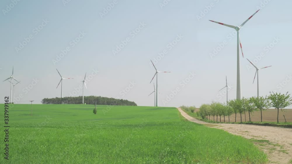 Wind turbines in the green fields in Germany. Powerful wind spinning the turbines. Green fields stretch to the horizon. The sky is hazy and grayish. A narrow dirt road goes through the field. Wind turbines in the green fields in Germany. Powerful wind spinning the turbines. Green fields stretch to the horizon. The sky is hazy and grayish. A narrow dirt road goes through the field.