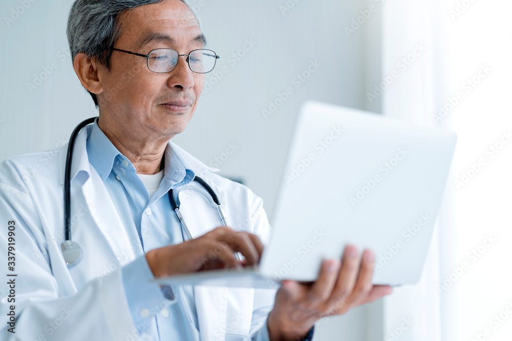 Senior asian male Doctor in hospital writing medical documentation and using laptop for manage white clinic background Senior asian male Doctor in hospital writing medical documentation and using laptop for manage white clinic background