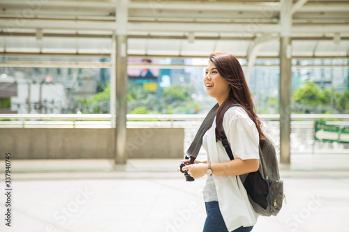 Portrait smiled young Asian woman in casual white shirt carrying a backpack walking on townsquare holding a camera, she is exiting while traveling photo