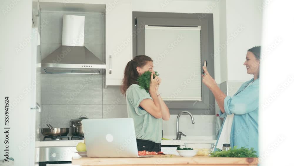 Two young pretty women making funny photos with a smartphone while cooking in the bright kitchen. Friends are having fun while cooking together. Two young pretty women making funny photos with a smartphone while cooking in the bright kitchen. Friends are having fun while cooking together.