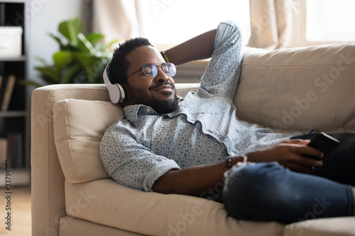 Tranquil carefree young african american man lying on comfy couch, wearing modern wireless headphones, listening to favorite classic music online, feeling peaceful mindful alone in living room. photo