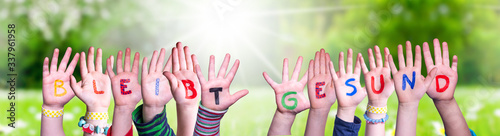 Kids Hands Holding Colorful German Word Bleibt Gesund Means Stay Healthy. Sunny Green Grass Meadow As Background