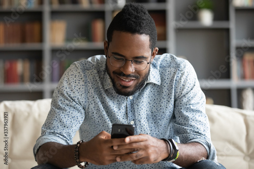 Smiling young african american man in glasses sitting on cozy couch, typing message on smartphone for friends or beloved woman girlfriend. Happy biracial guy playing online game on mobile phone.