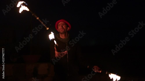 Professional juggler performing juggling tricks with 3 fire clubs, lighting the outdoor dark night scene. Slow motion medium close up shot.