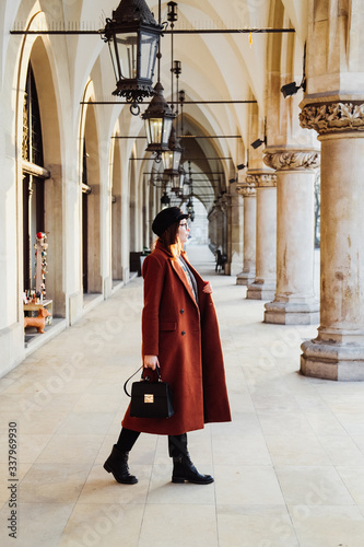 A beautiful young woman in a terracotta coat walks through the old city.