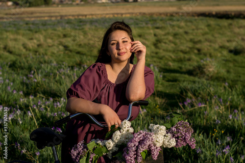 A beautiful young Caucasian smiling happy girl in purple dress in a wreath and with a bicycle a basket of blooming lilac in the countryside. Model plus size. Spring, rustic style. Circlet of flowers
