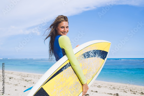 Adult lady in wetsuit with surf board standing and looking at camera on ocean shore in sunny day