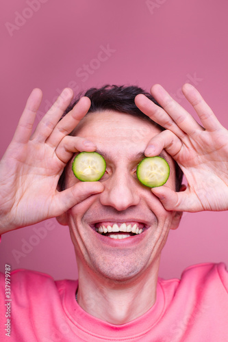 Vertical portrait of weird strange funny guy smiling. Hold cucumber slices in front eyes. Facial treatment and care. Isolated over pink background.