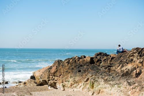 Rear view of pensive, depressed man sitting on coast rock, looking at sea horizon.Copy space, unrecognizable person