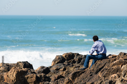 Rear  view of pensive, depressed man sitting on coast rock, looking at sea horizon.Copy space, unrecognizable person, lens flare, sunset light © Avatar_023