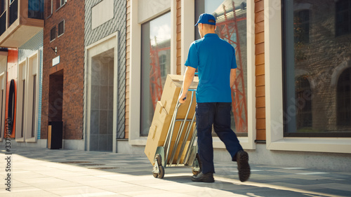 Delivery Man Pushes Hand Truck Trolley Full of Cardboard Boxes, Packages For Delivery. Professional Courier Working Efficiently, Quickly. Walks Through Stylish Modern Urban Area. Back View  photo