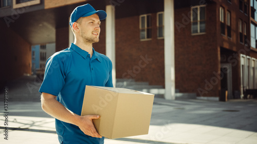 Handsome Delivery Man Holds Cardboard Box Package Walks Through Modern Stylish Business District. Courier On the Way to Deliver Postal Parcel to a Client. © Gorodenkoff